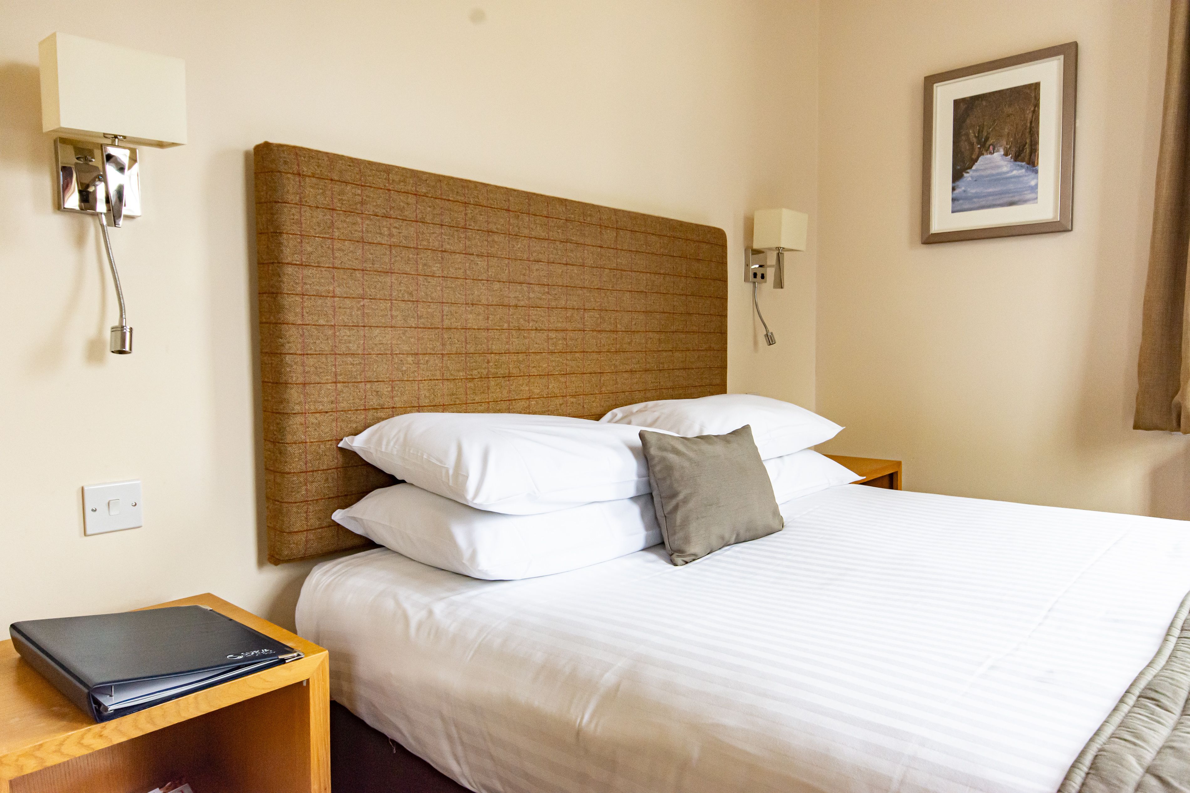 A neatly made bed with white linens and a single gray pillow, a wooden nightstand with a laptop, a wall-mounted light fixture, and a framed picture on the wall.