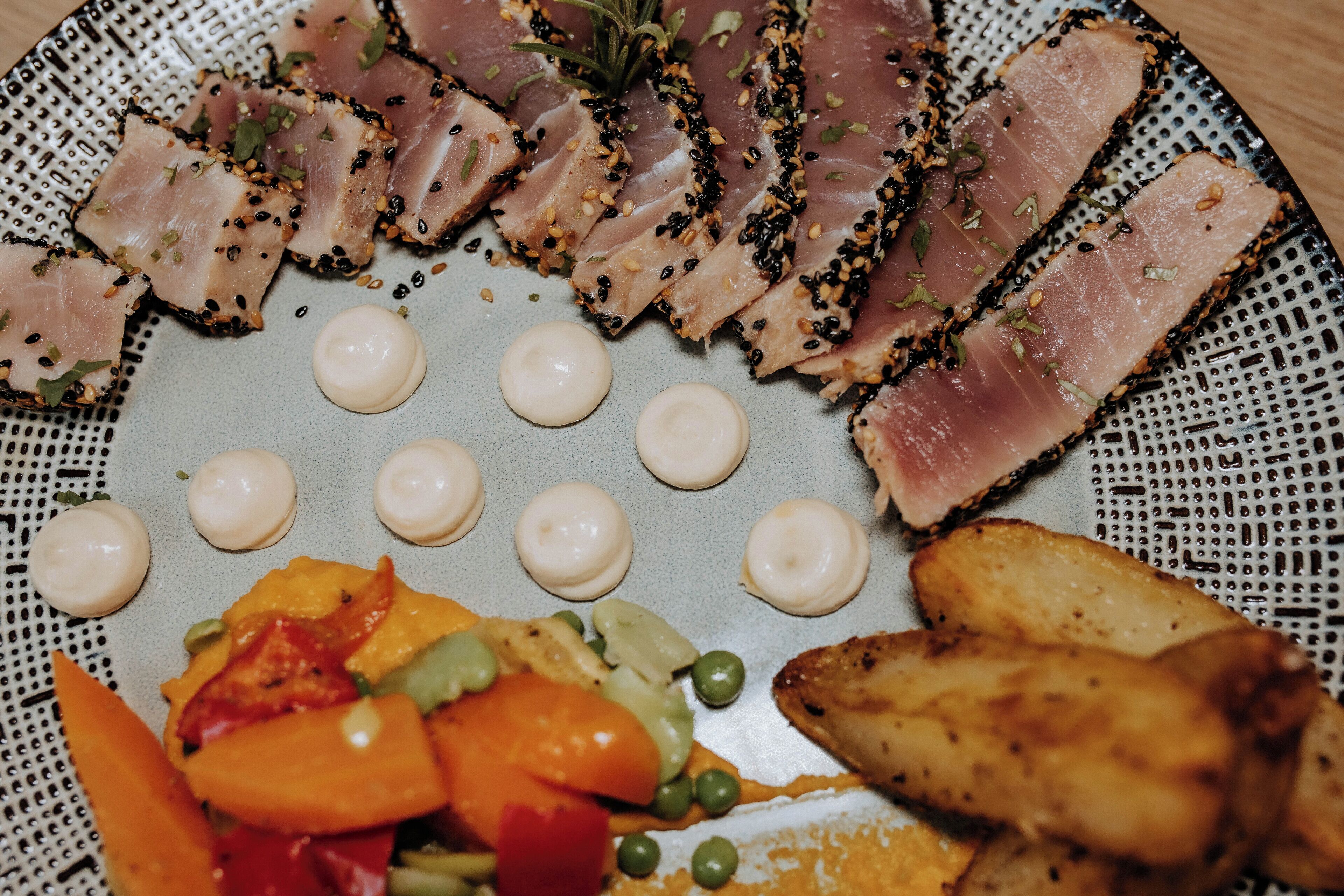 A plate of sliced meat with sesame seeds, garnished with herbs, accompanied by roasted vegetables and small round items.