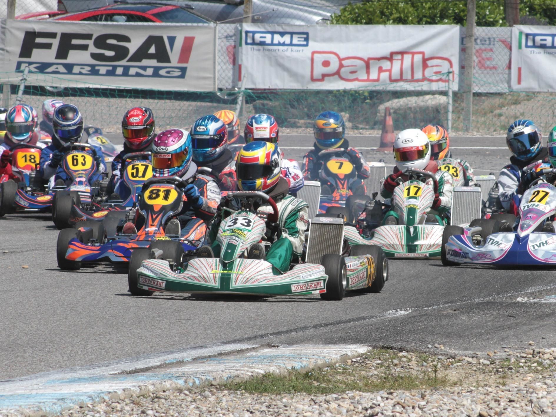 A go-kart racing event with multiple participants in colorful racing suits and helmets, racing on a track with visible sponsor banners in the background.