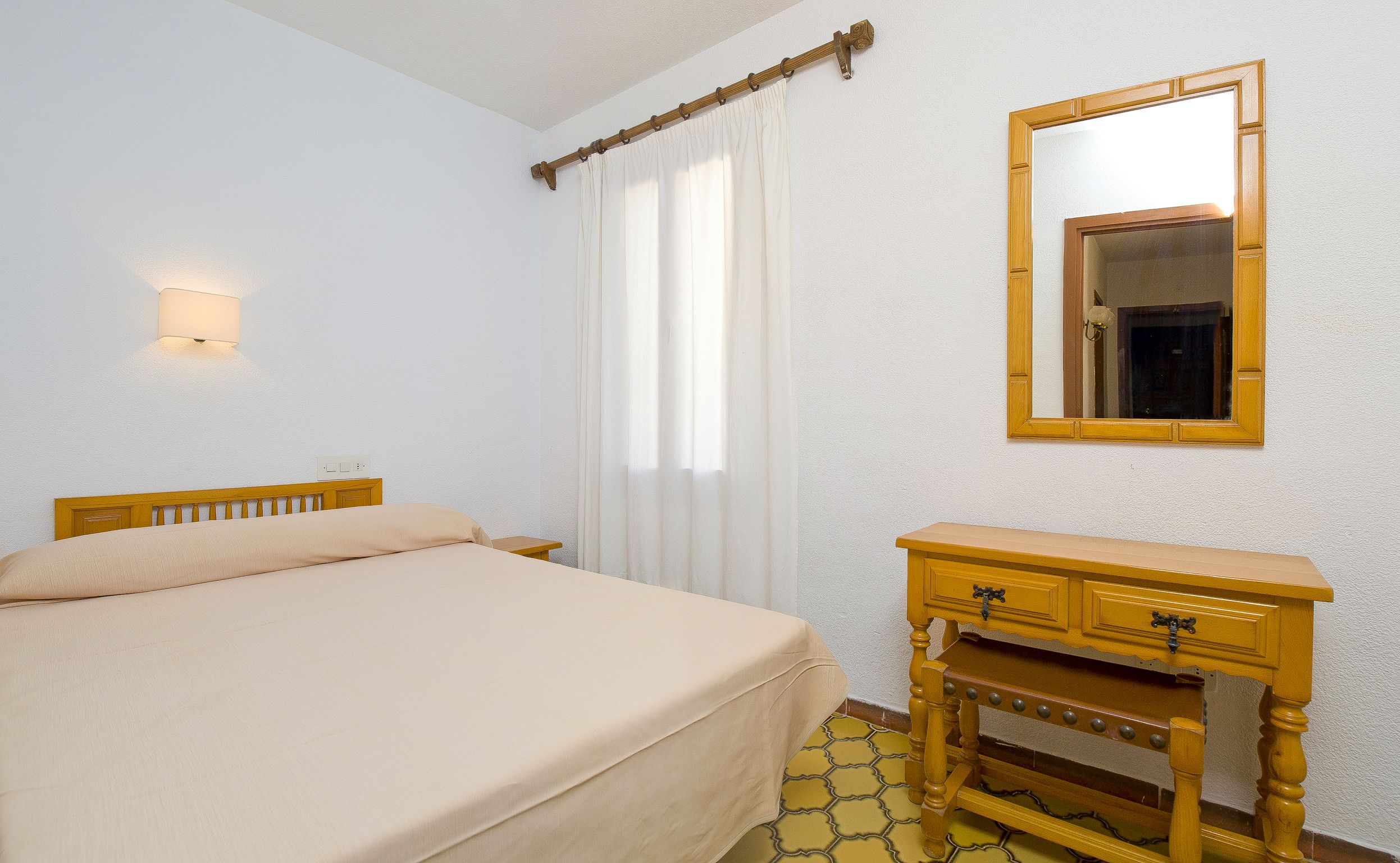 A bedroom with a bed, a wooden dresser, a mirror, and a wall-mounted light.