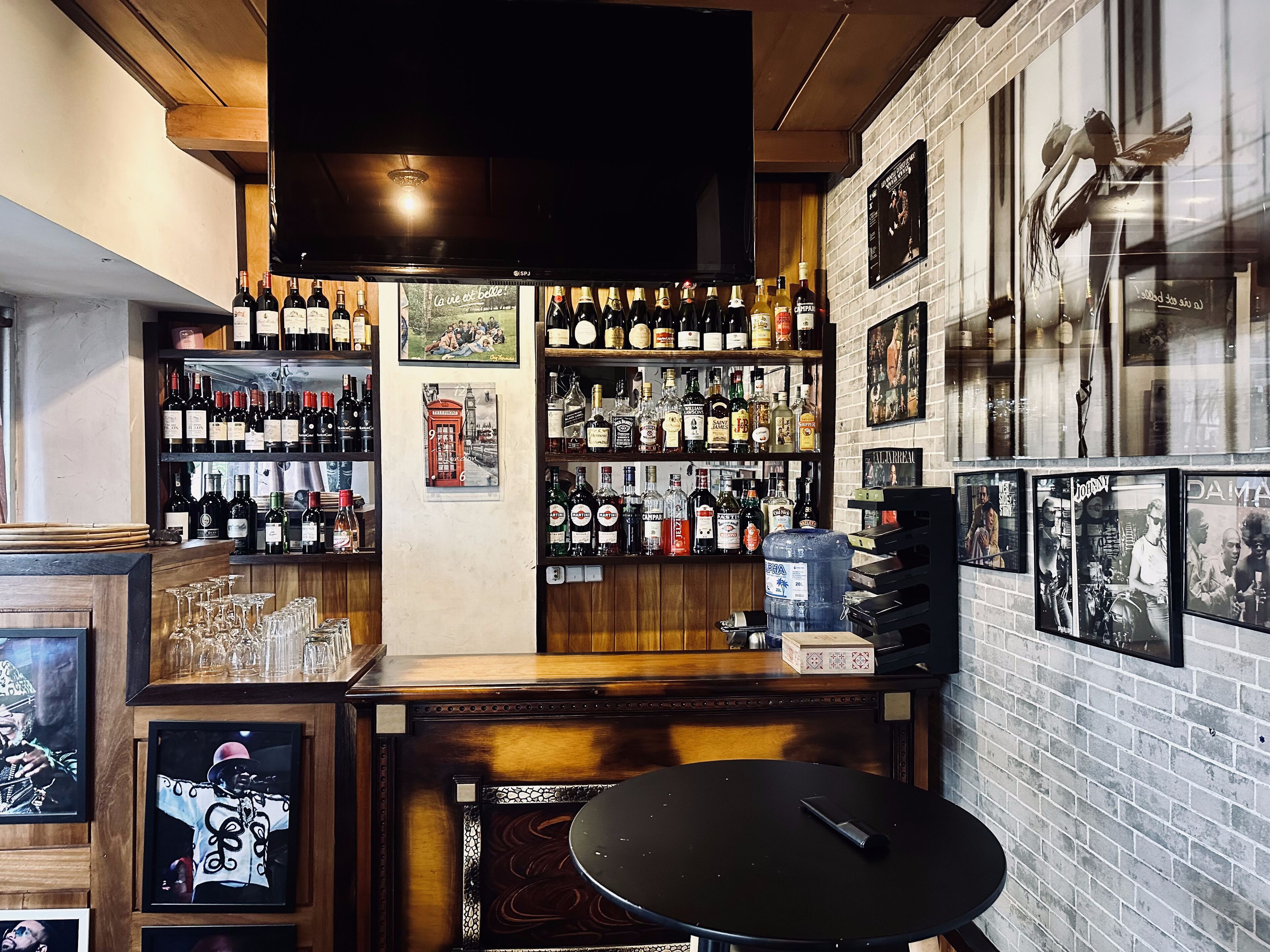 A bar with wooden shelves stocked with various bottles, a large water dispenser, and framed pictures on the walls.