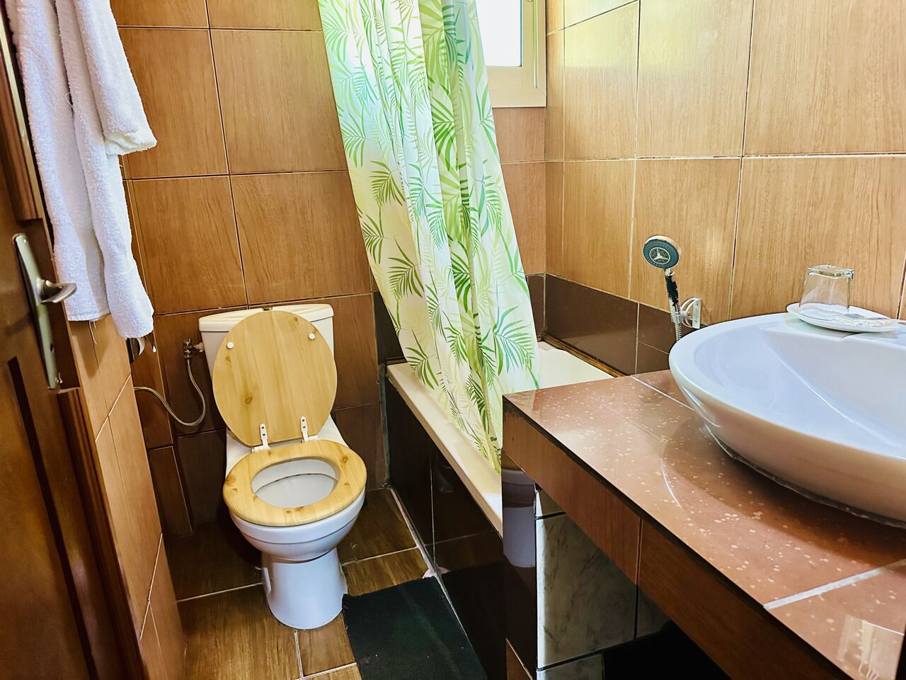 Une salle de bain avec des toilettes en bois, un lavabo doté d’un plan de travail en marbre et un rideau de douche orné d’un motif de feuilles.