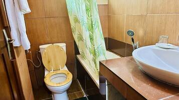 Un baño con inodoro de madera, lavabo con encimera de mármol y cortina de ducha con estampado de hojas.