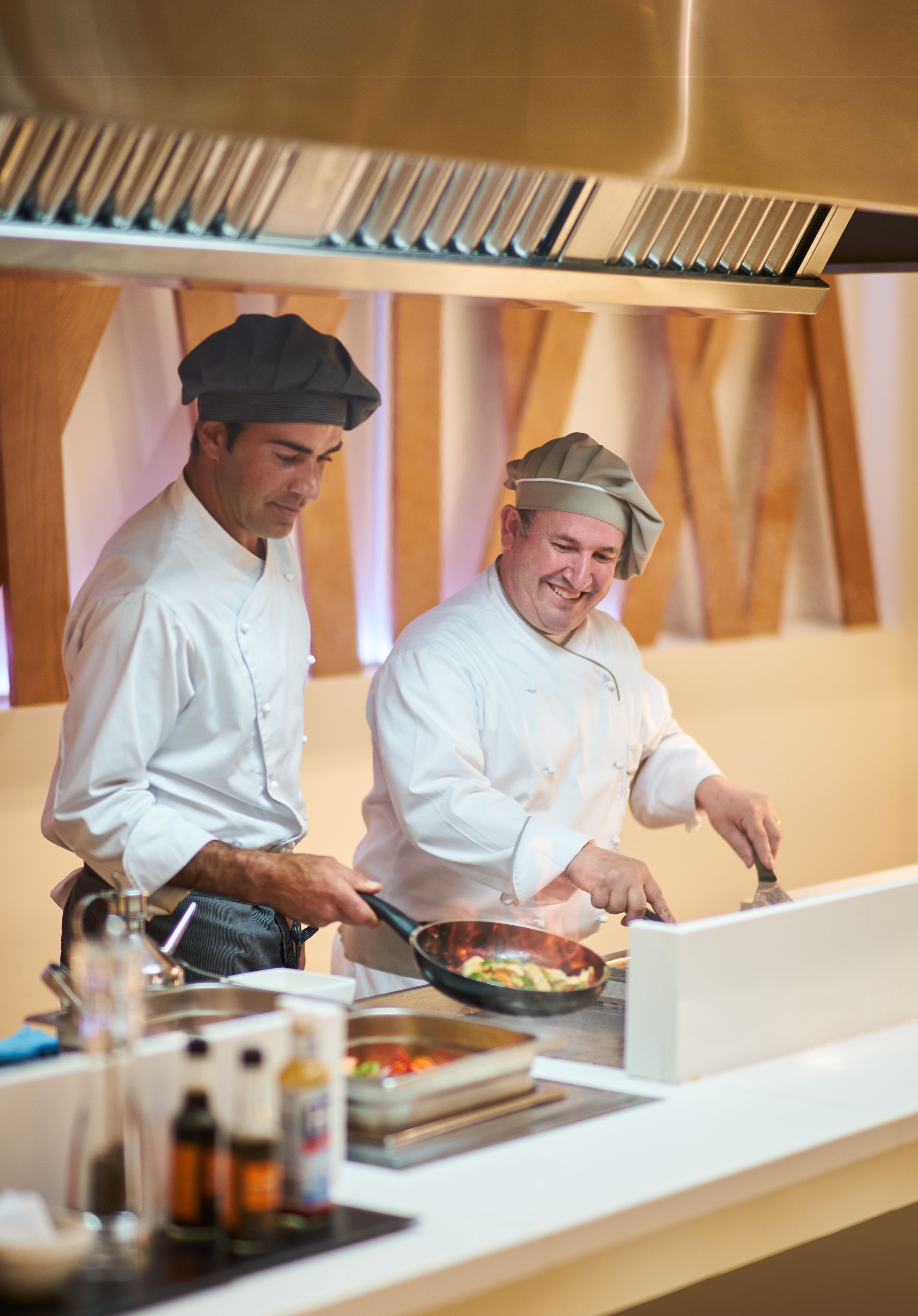 Two chefs in a professional kitchen with stainless steel appliances and wooden accents.