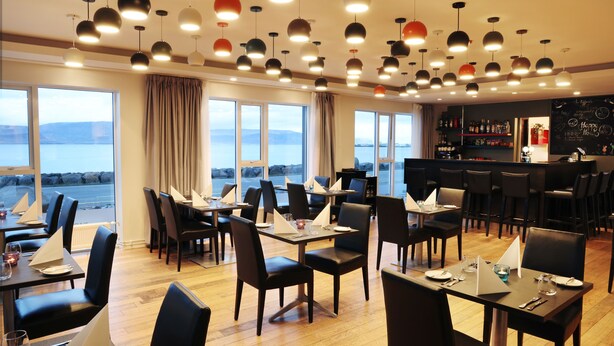 A restaurant with multiple tables and chairs, large windows with a view of the sea, and a bar area with shelves of bottles. - Fosshotel Westfjords (Vesturbyggð)