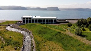 A modern building with a glass facade, situated by a body of water with a rocky shoreline and grassy terrain. - Fosshotel Westfjords (Vesturbyggð)