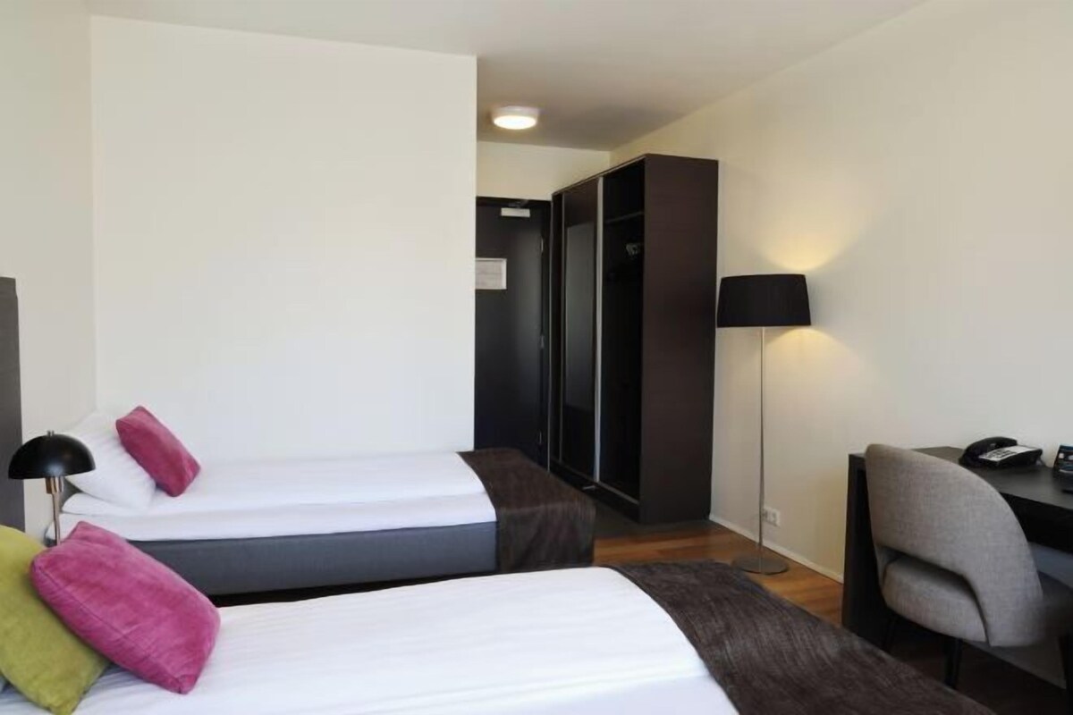 A hotel room with two beds, a desk, a chair, a lamp, and a wardrobe.