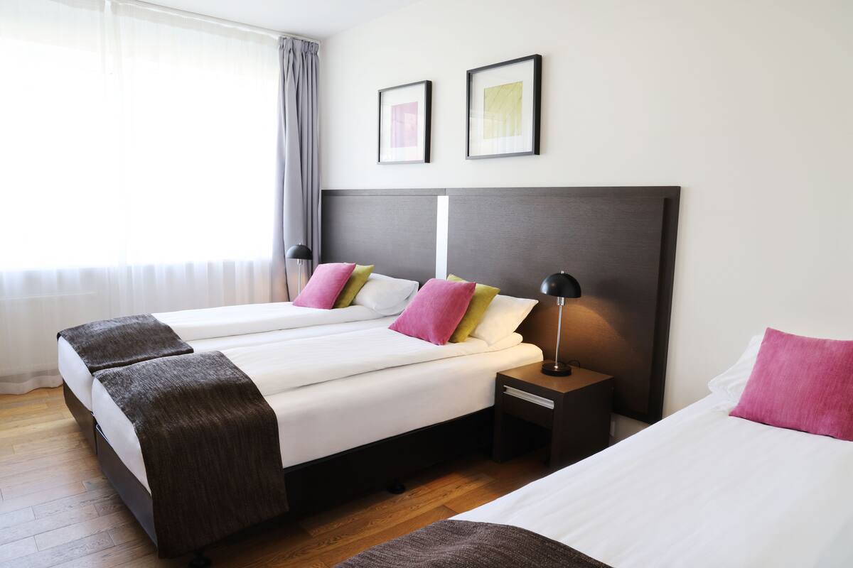 Ein Hotelzimmer mit zwei Betten, einem dunklen Kopfteil, einem Nachttisch mit Lampe und einem Fenster mit Vorhängen.