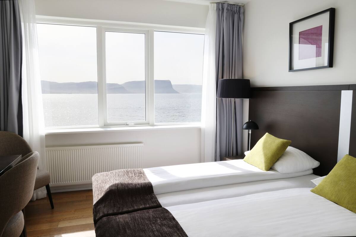 Ein Hotelzimmer mit großem Fenster und Meerblick, einem Bett mit weißer Bettwäsche und zwei Kissen, einem Nachttisch und einer Lampe.