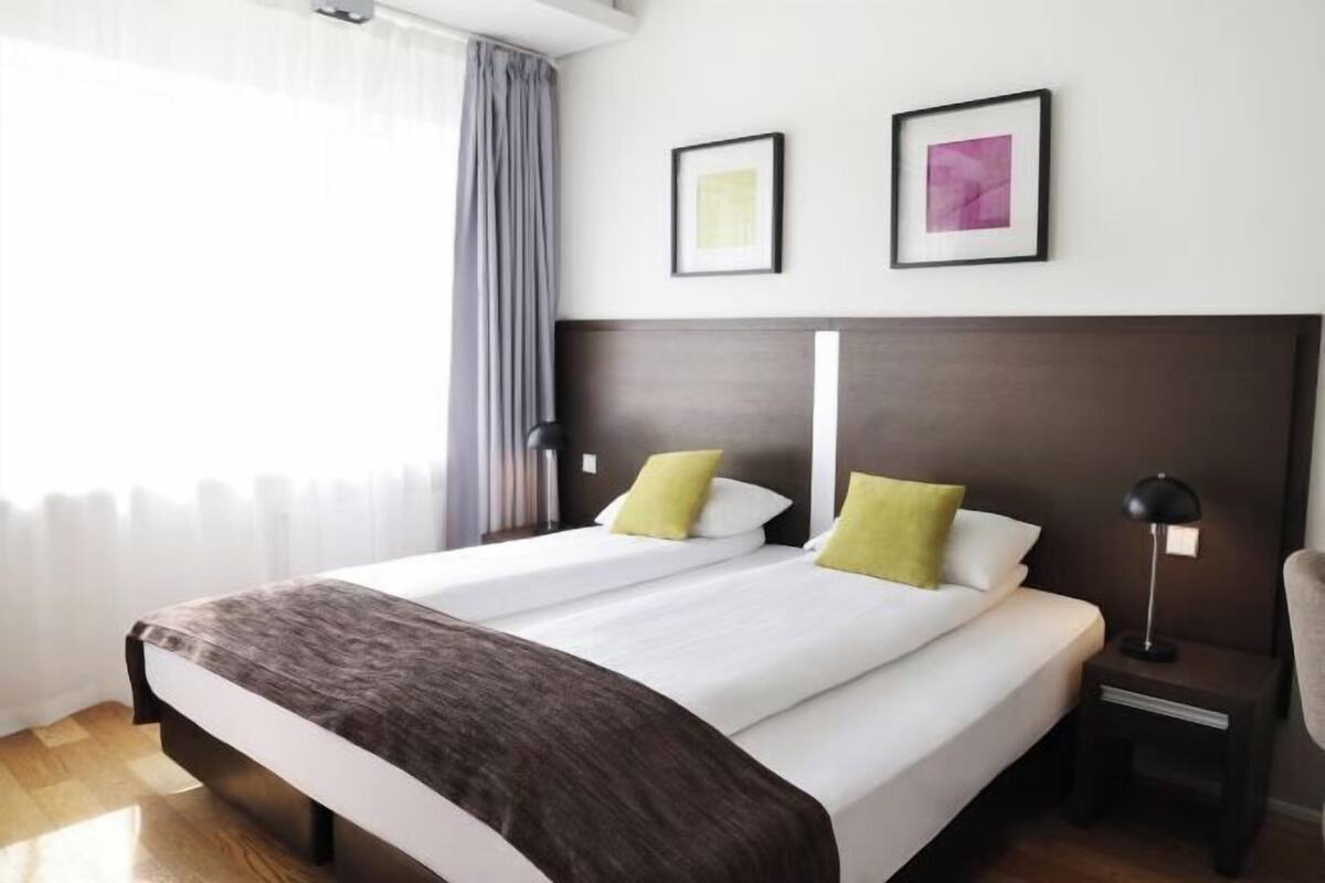 Ein Hotelzimmer mit einem großen Bett, zwei Kissen, einem Nachttisch mit Lampe und zwei gerahmten Bildern an der Wand.