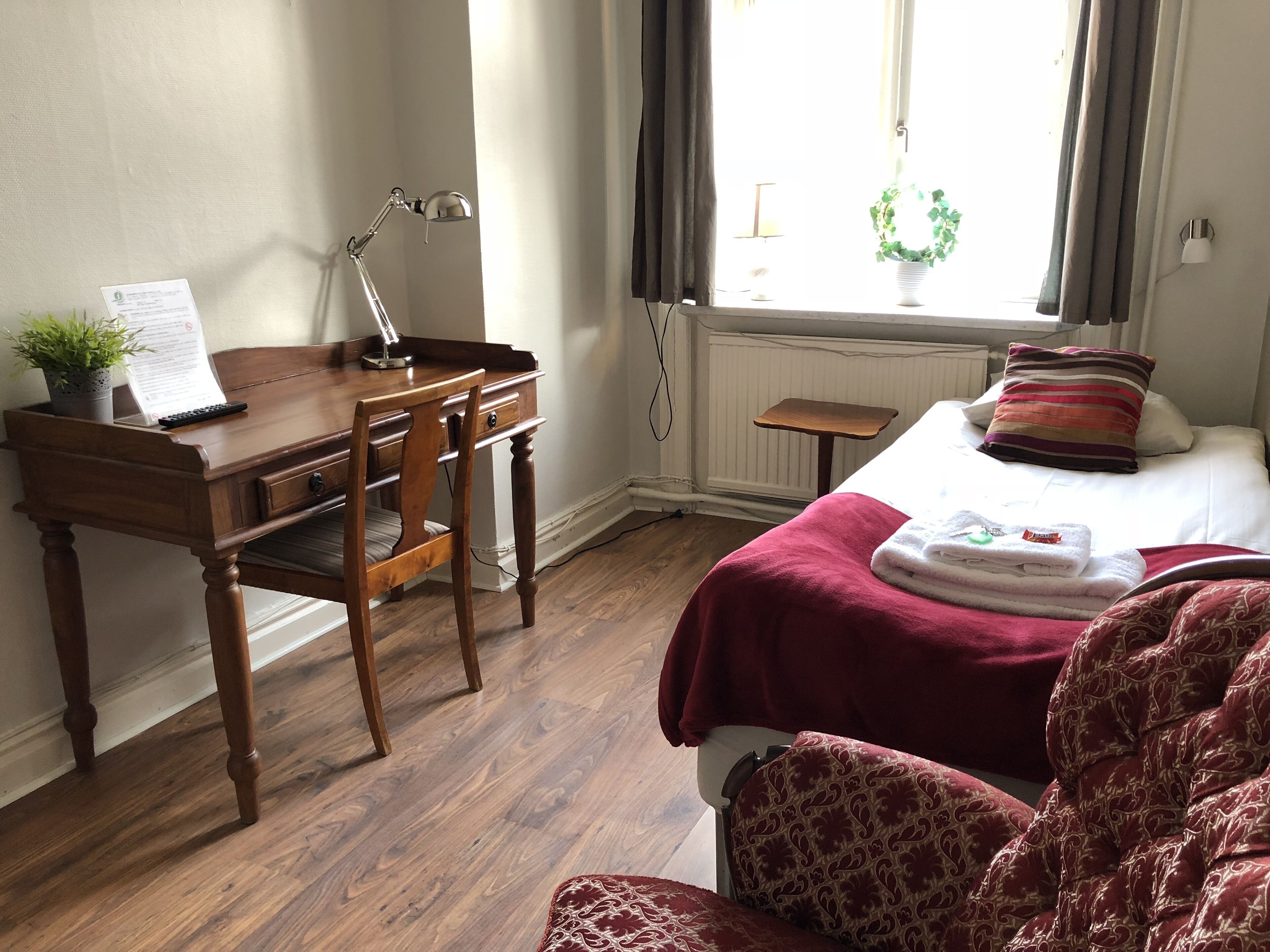 A hotel room with a wooden desk, chair, and a bed with a red blanket. There is a window with curtains and a plant on the windowsill.