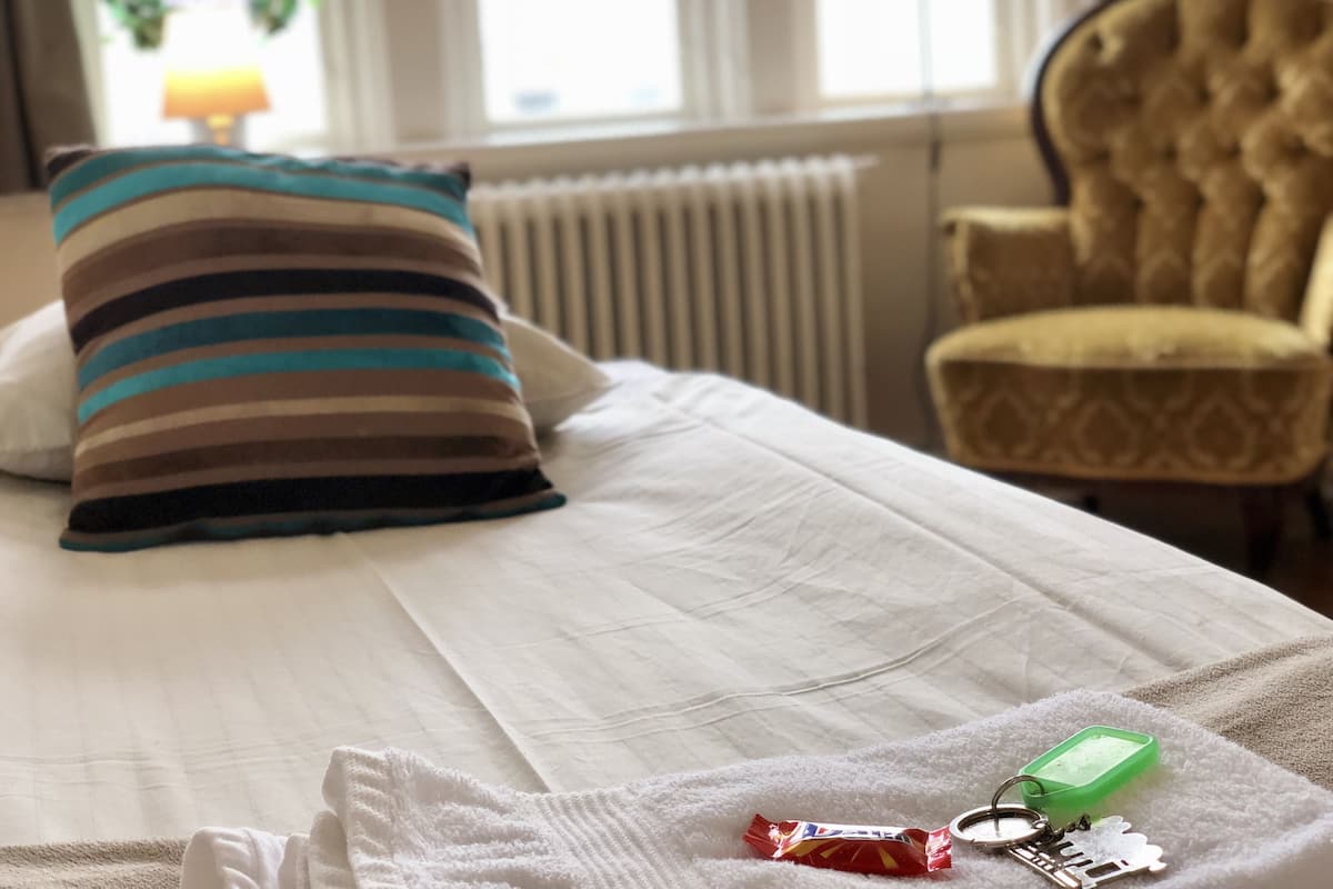 A hotel room with a bed, a striped pillow, a chair, and a small table with a green container and a red item on it.