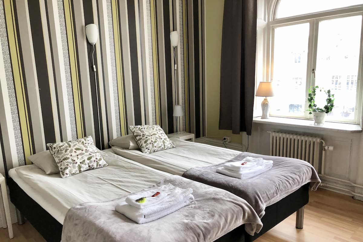 A hotel room with two beds, striped wallpaper, a window with a view, and a radiator.