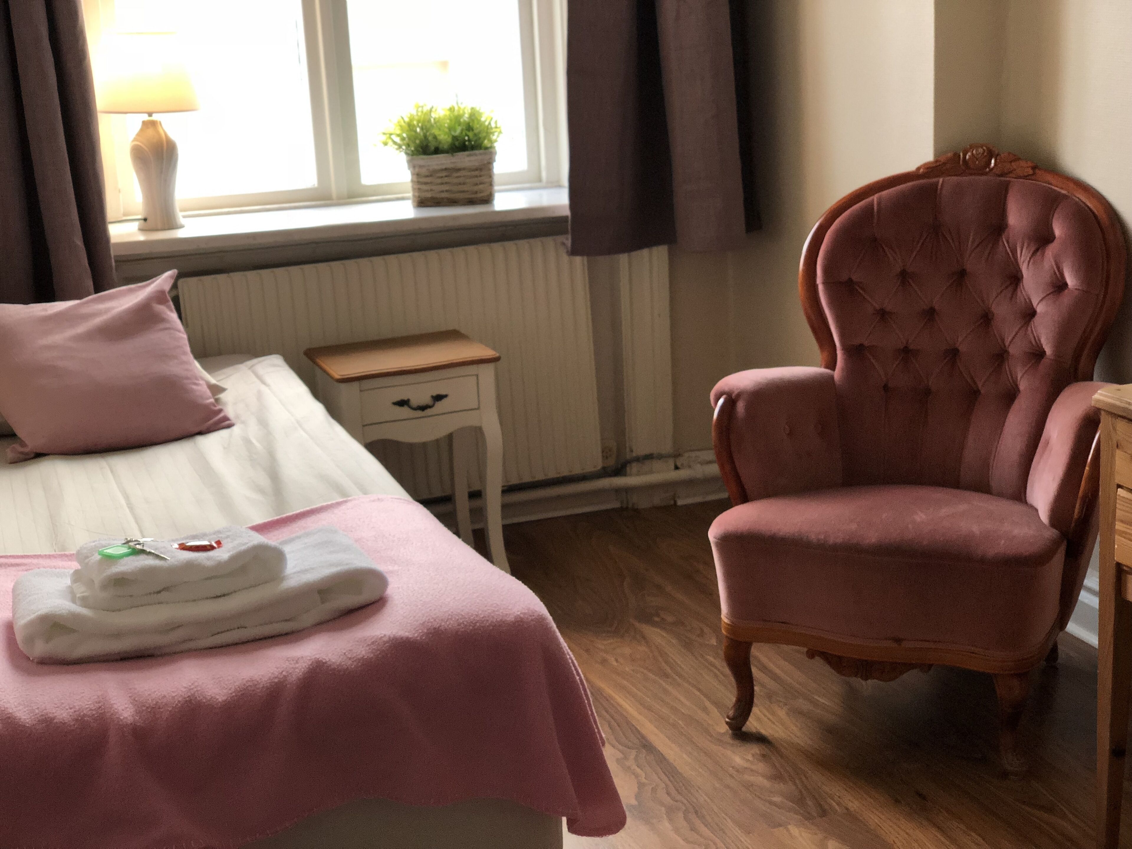 A bedroom with a bed, a pink chair, a bedside table, a lamp, and a window with a plant.