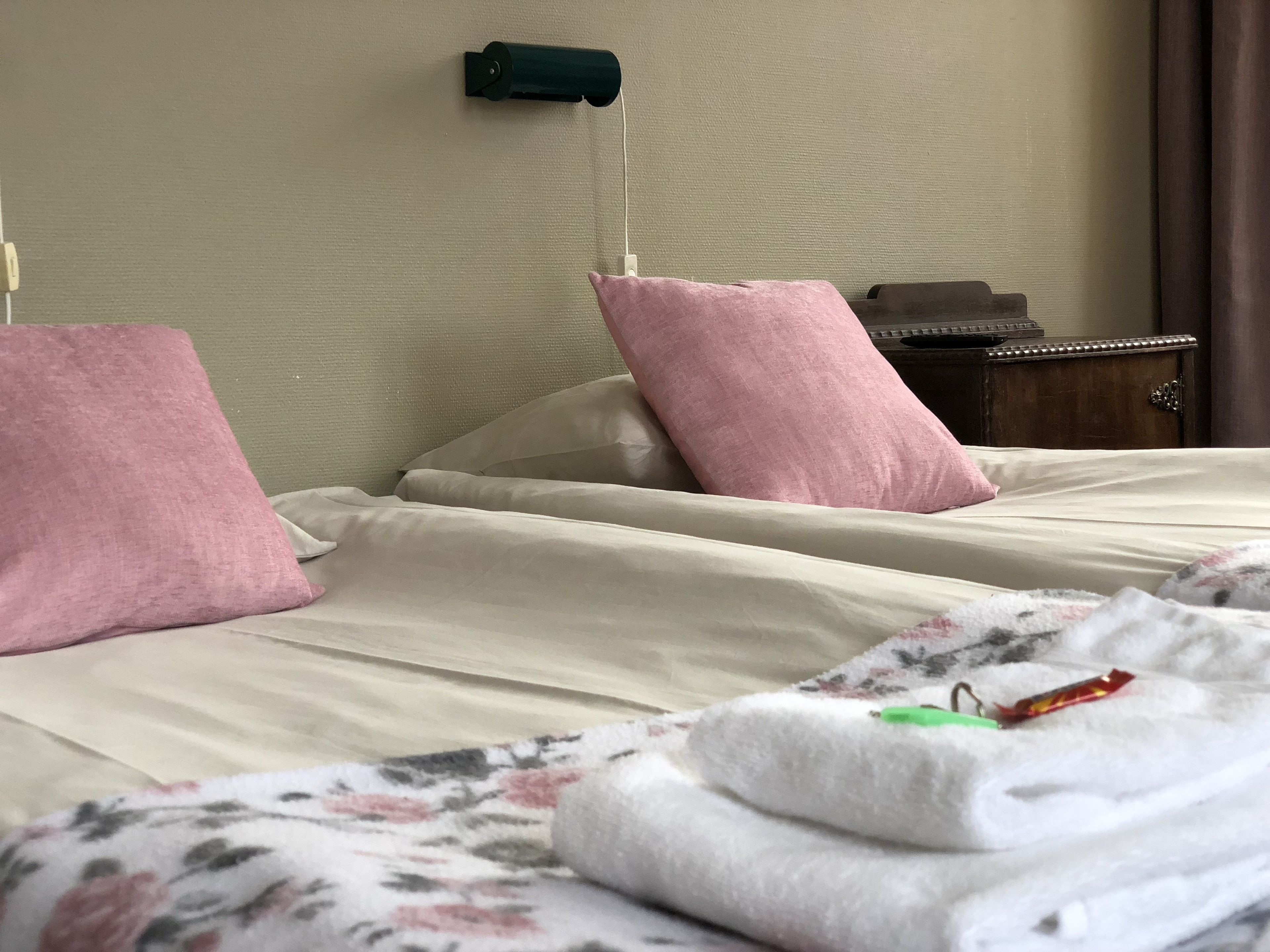 Ett hotellrum med två sängar, var och en med en rosa kudde och ett beige sängöverkast. Det finns ett träbord vid sängen med en lampa och ett litet bord.