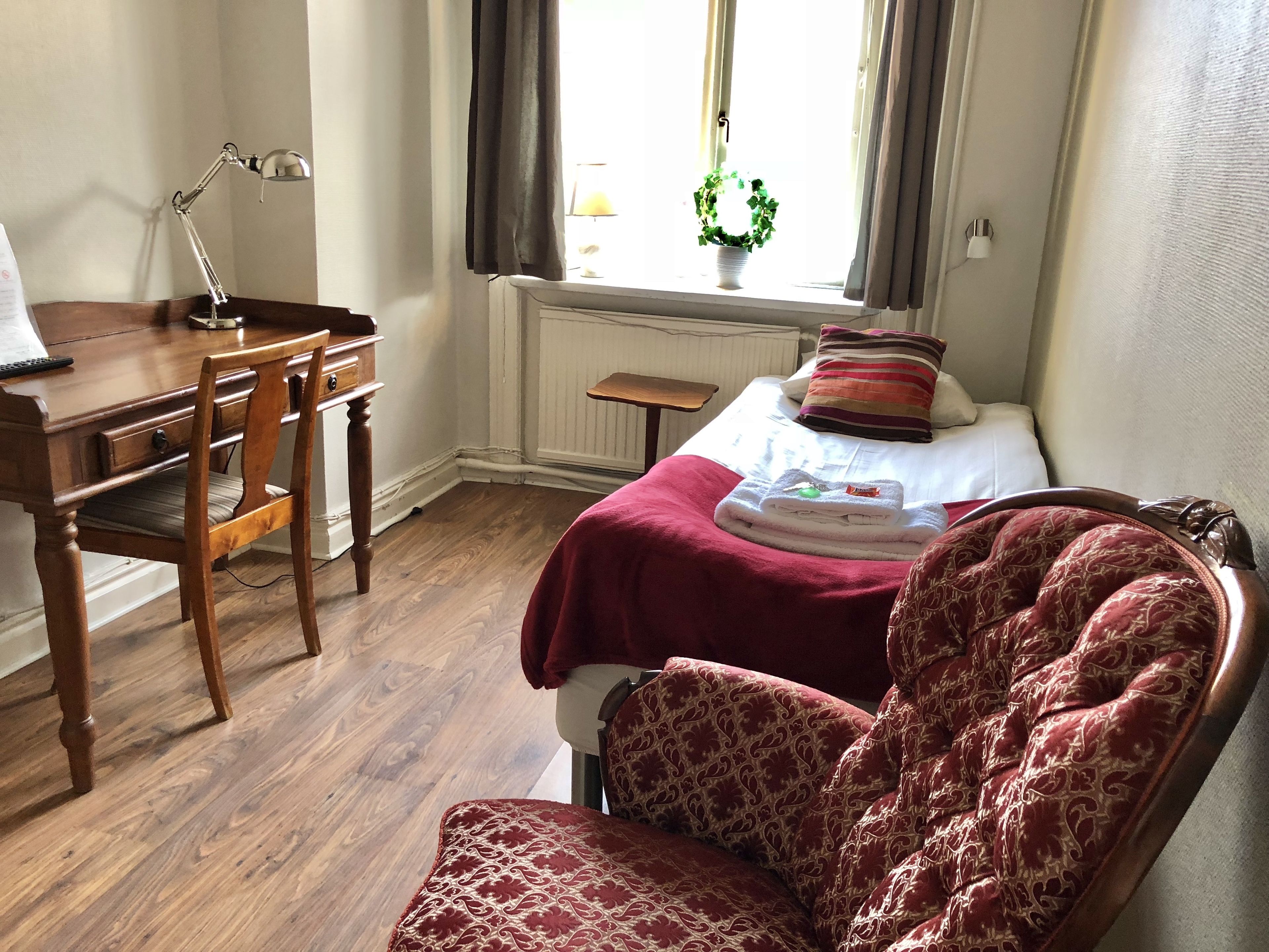 Une chambre avec un bureau en bois, une chaise, un lit recouvert d’une couverture rouge et une fenêtre avec des rideaux.