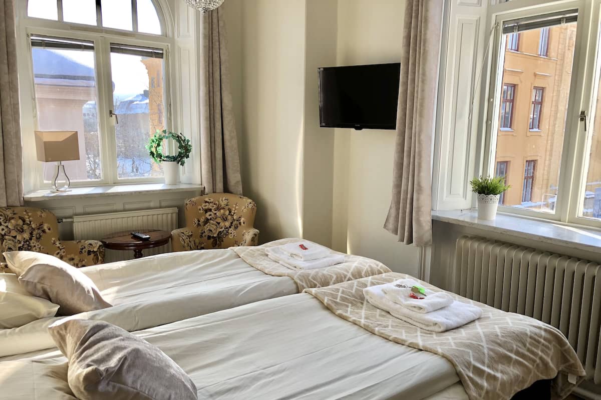 A hotel room with two beds, a TV, a window with curtains, and a view of buildings outside.
