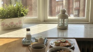 Una mesa de desayuno con un plato de comida, una taza, una vela y una planta en maceta junto a una ventana que ofrece vistas de un edificio de ladrillo.