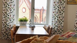 Un comedor con una mesa de madera, sillas, un cuenco de pan, una planta en maceta y una ventana con cortinas florales.