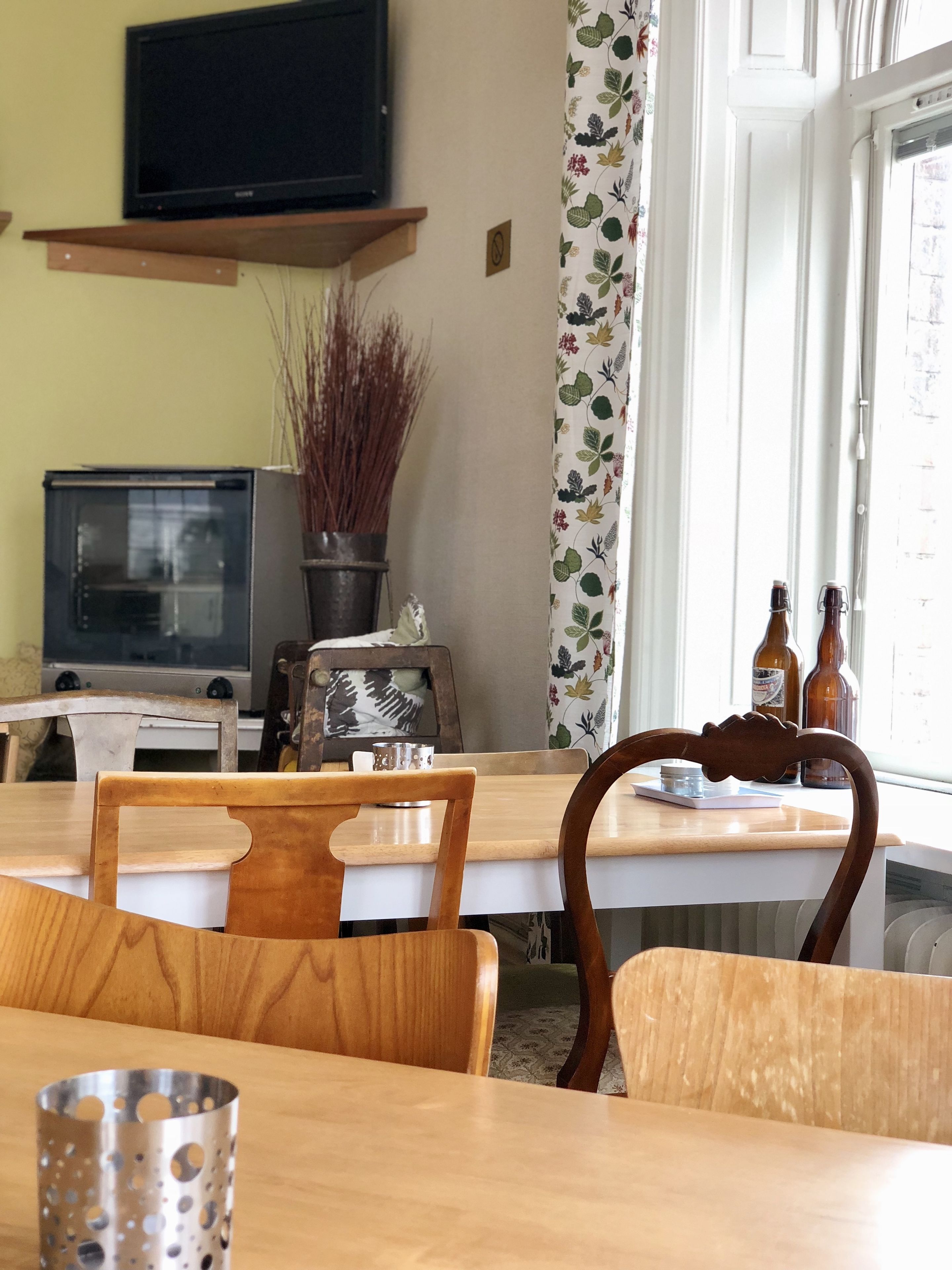 Un comedor con sillas de madera y una mesa, un televisor en un estante, y un ventanal con cortinas.