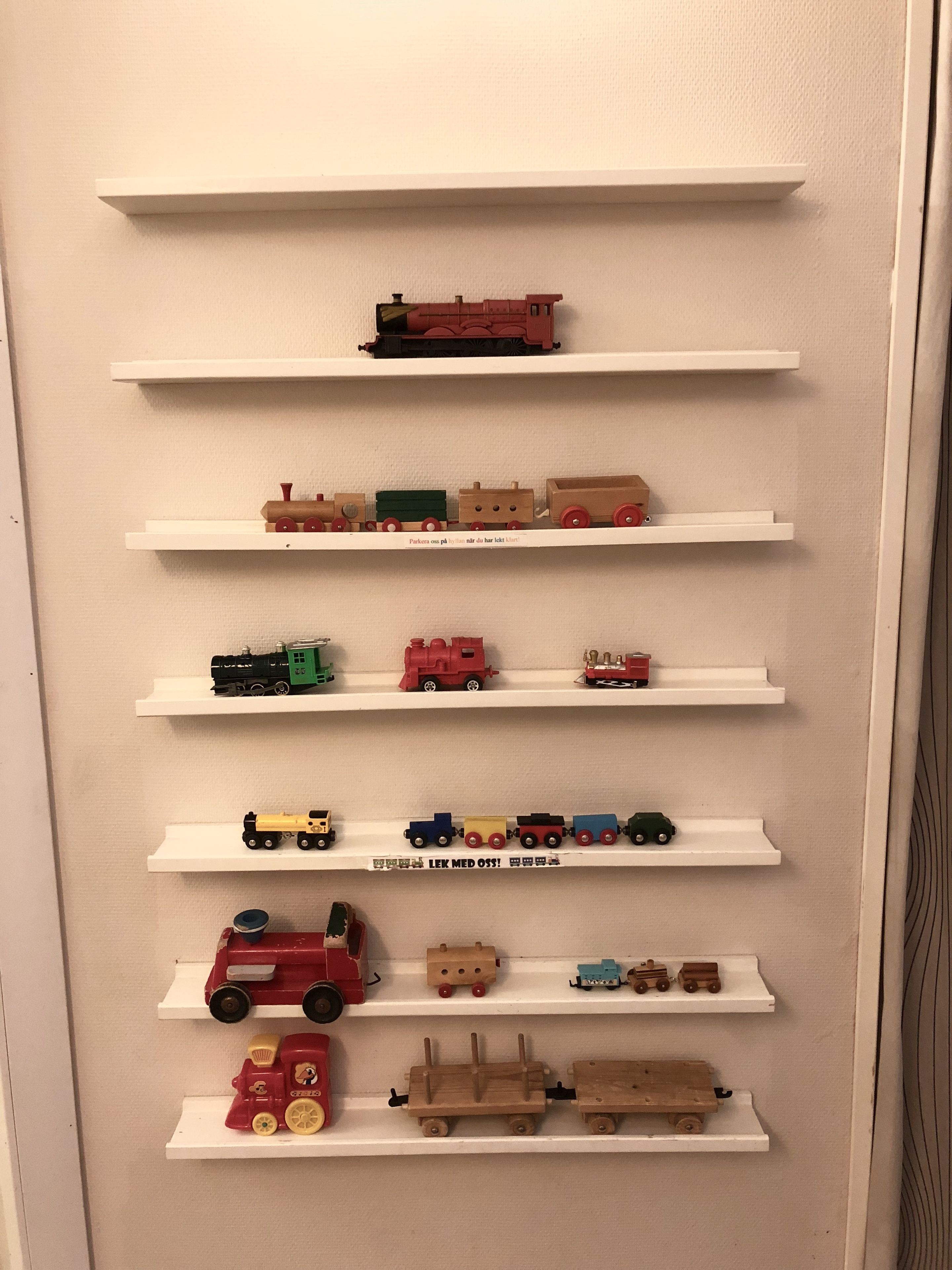 Una pared con varias repisas blancas que exhiben diversos trenes de juguete y vehículos.