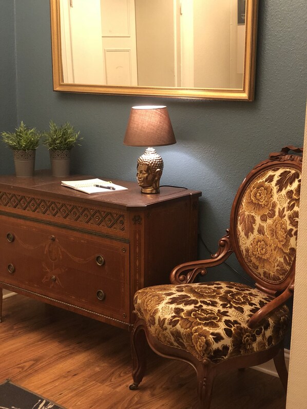 Una habitación con un aparador de madera, una silla con tapicería floral, una lámpara con la figura de Buda y un espejo en la pared.