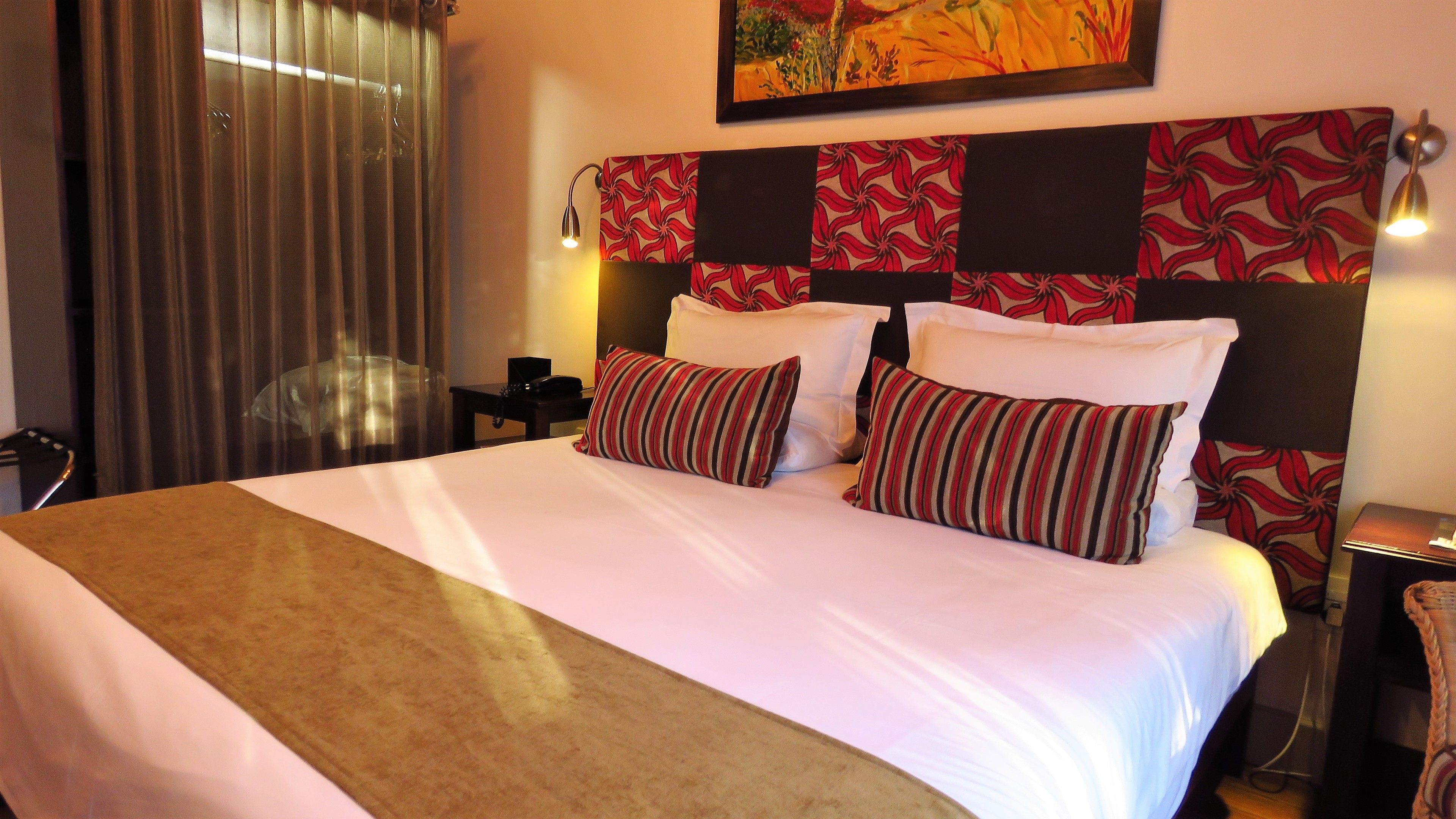 A hotel room with a large bed, a headboard with a floral pattern, a bedside table with a lamp, and a painting on the wall.