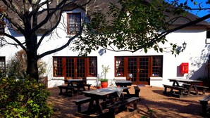 Un patio con mesas y bancos de picnic de madera, rodeado de plantas en macetas y un árbol grande.