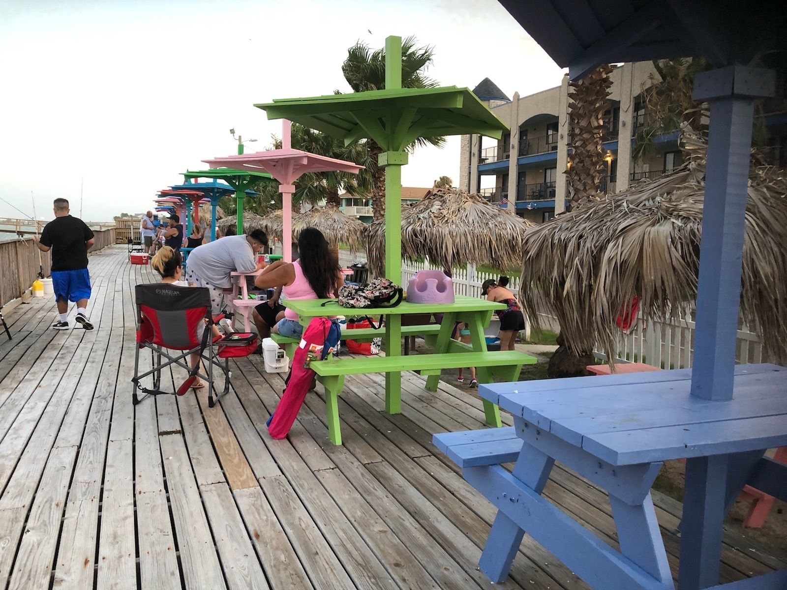 A wooden pier with picnic tables and umbrellas, overlooking a beach area with palm trees and a building in the background.