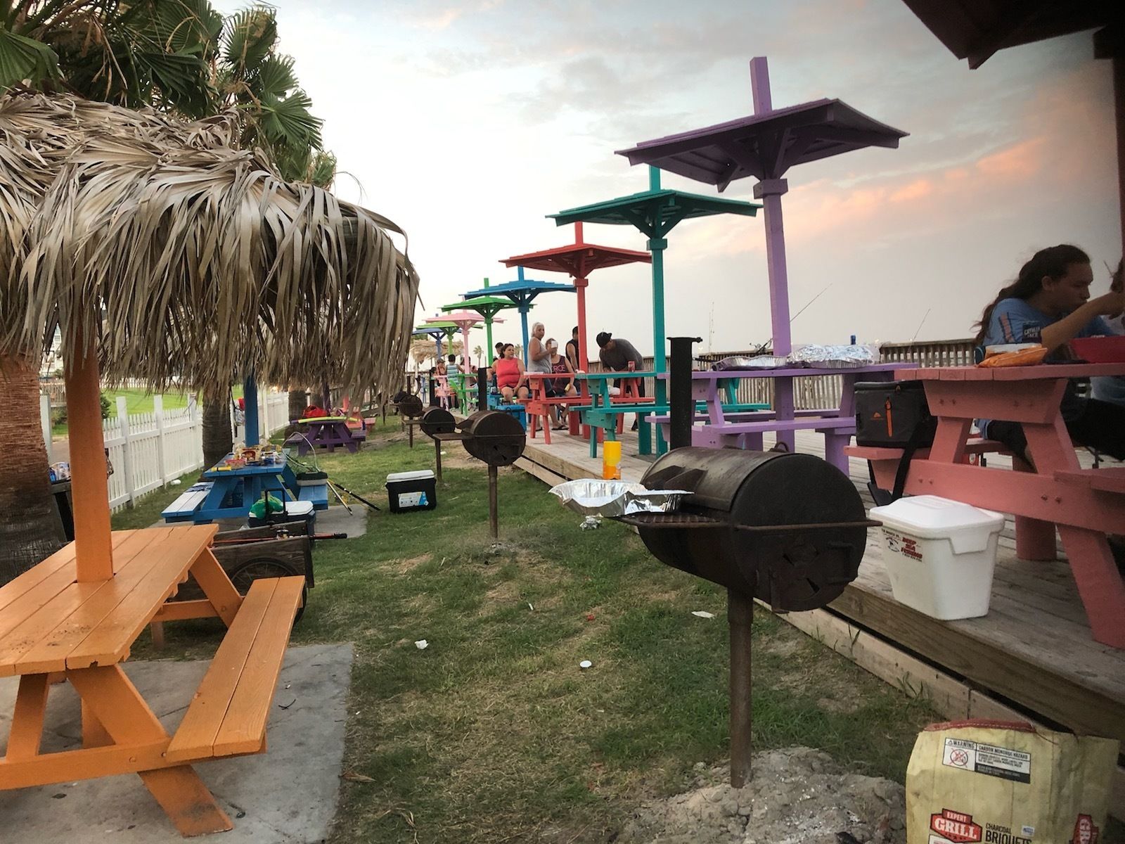 A beachside barbecue area with picnic tables, barbecue grills, and colorful umbrellas.