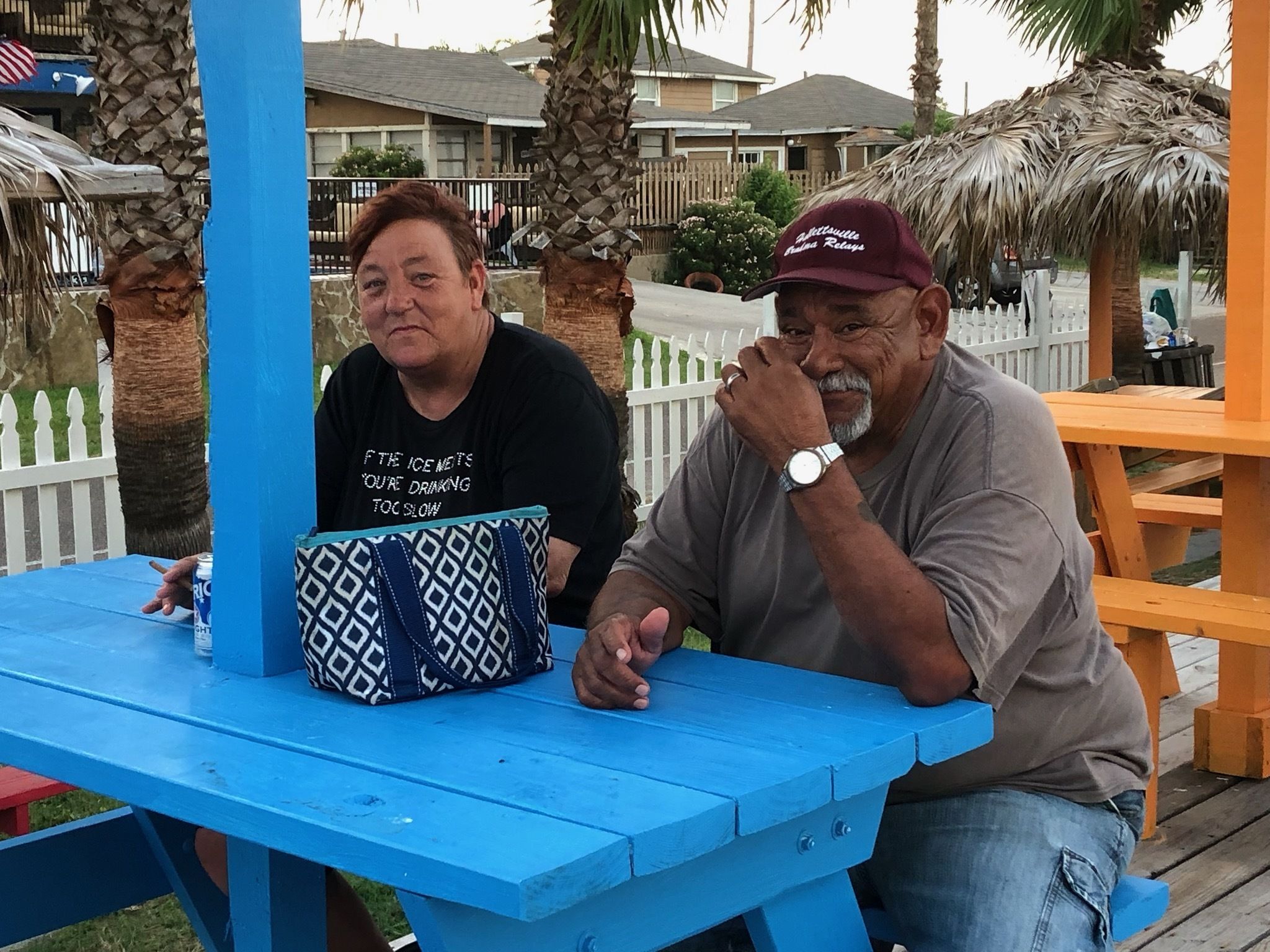 Two people sitting at a blue picnic table with a patterned bag on it.