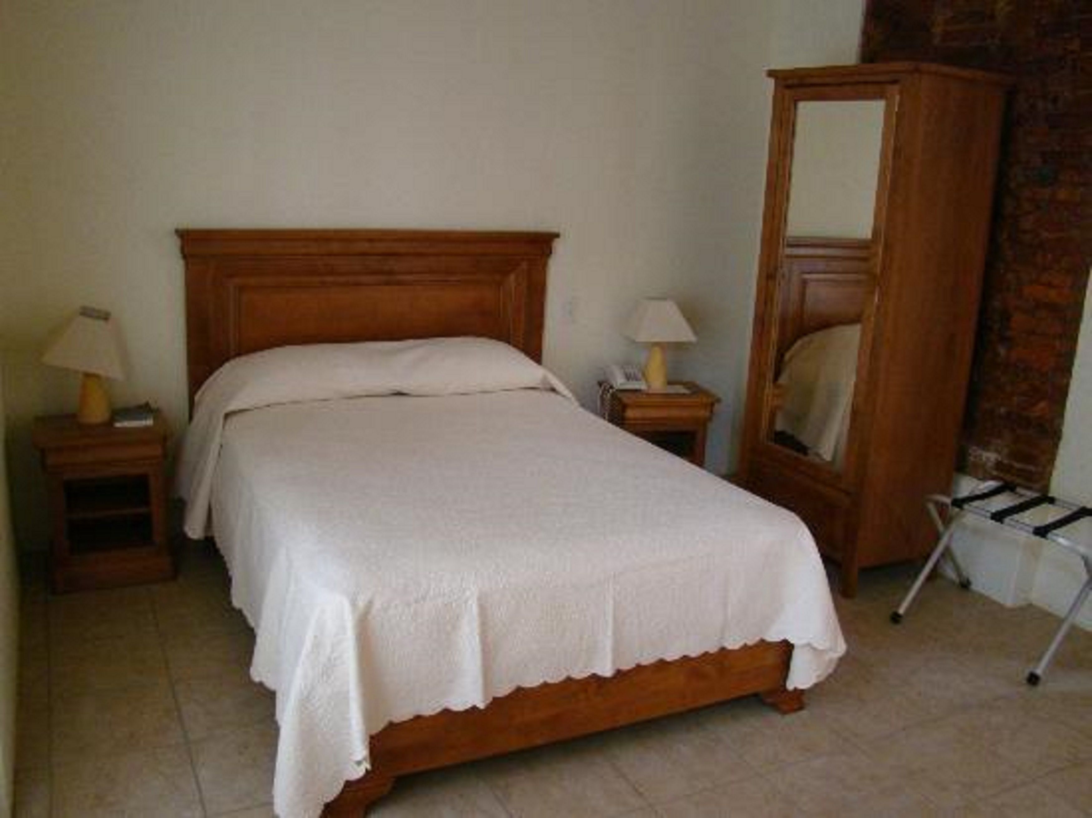 A bedroom with a wooden bed, two bedside tables with lamps, a tall mirror, and a brick wall.