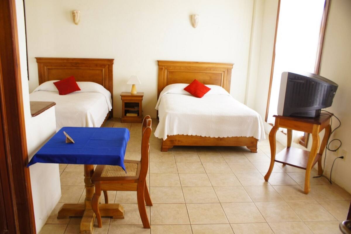 Une chambre d’hôtel avec deux lits, un téléviseur posé sur un meuble, une petite table recouverte d’une nappe bleue et une chaise.