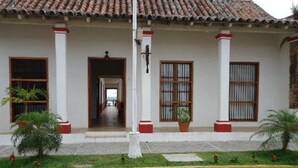Un edificio con techo de tejas, paredes blancas y una entrada central con puerta y ventanas. Hay plantas en macetas y una bordillo amarillo en la parte delantera.