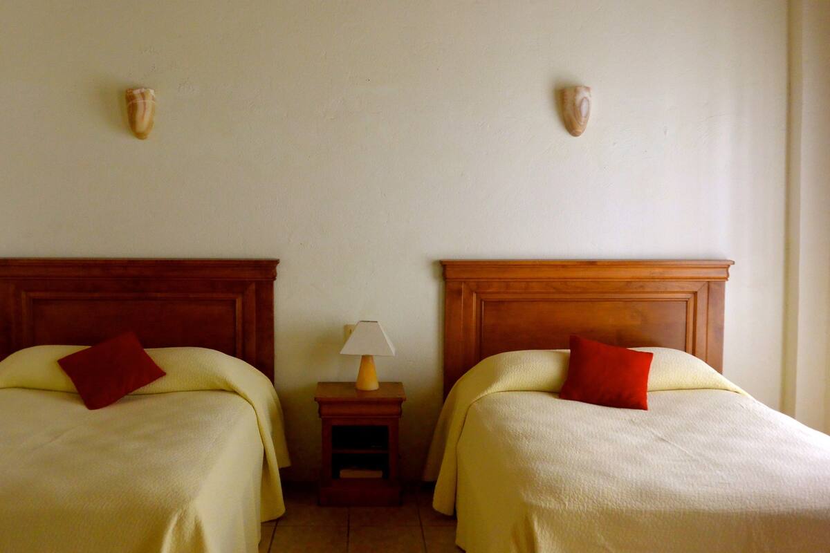 Deux lits simples avec des têtes de lit en bois, une table de chevet avec une lampe et des appliques murales décoratives.