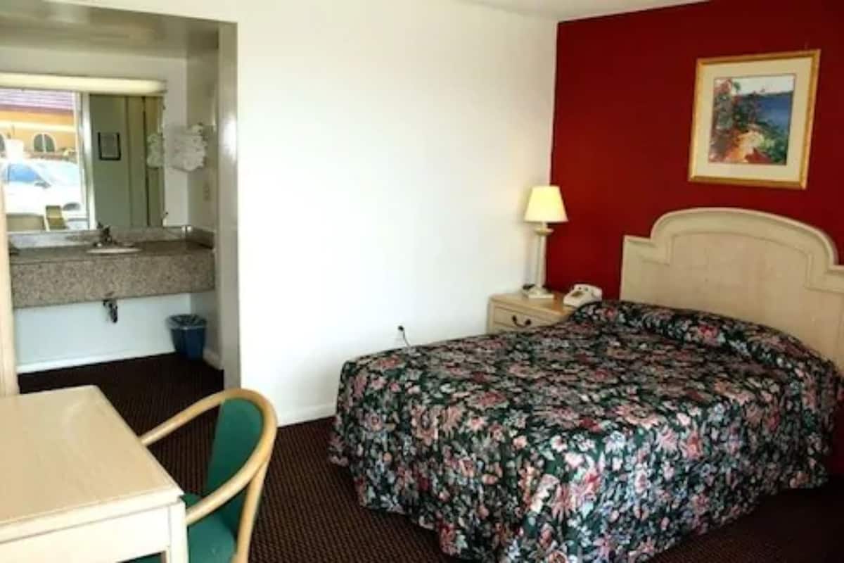 A hotel room with a bed, a desk, a chair, a nightstand, and a painting on the wall.