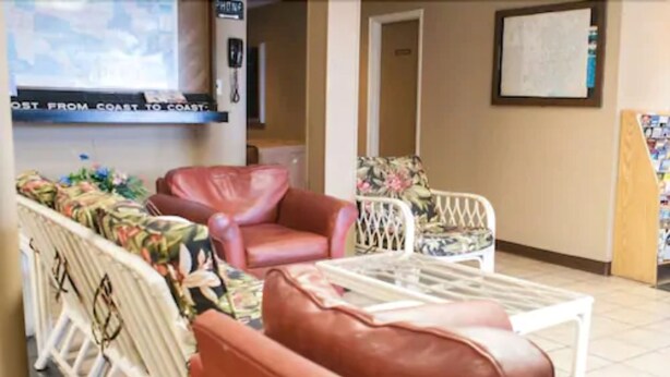 A lobby with a seating area featuring a brown leather sofa and a white wicker chair. A glass coffee table is in front of the sofa. - Scottish Inn Jacksonville Downtown (Jacksonville)