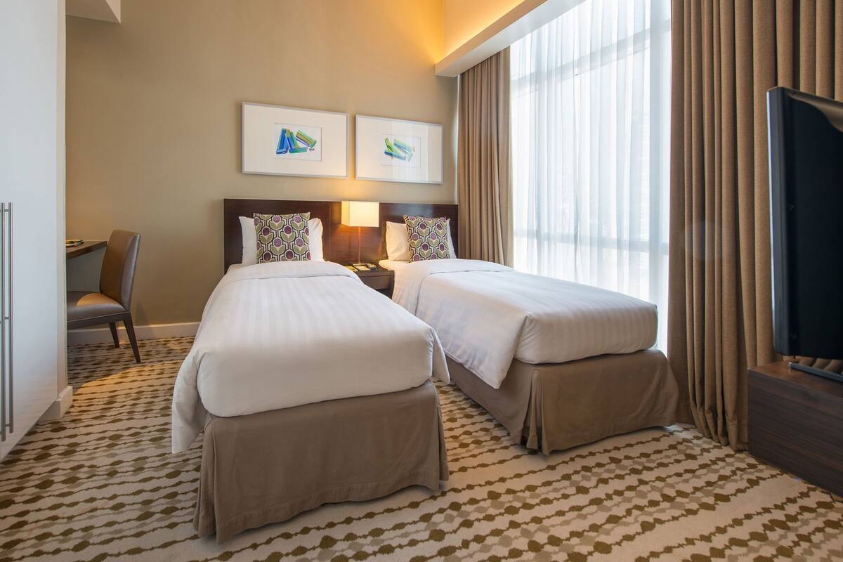 A hotel room with two beds, a desk, a chair, a TV, and artwork on the wall.