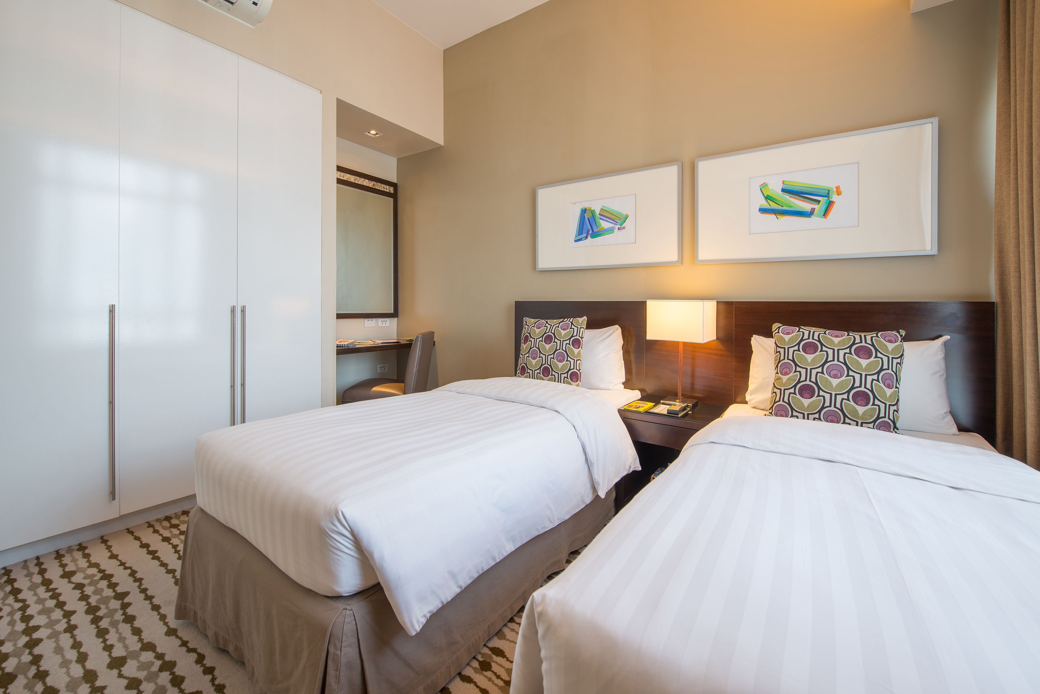 Habitación de hotel con dos camas, un armario, un escritorio y dos cuadros en la pared.