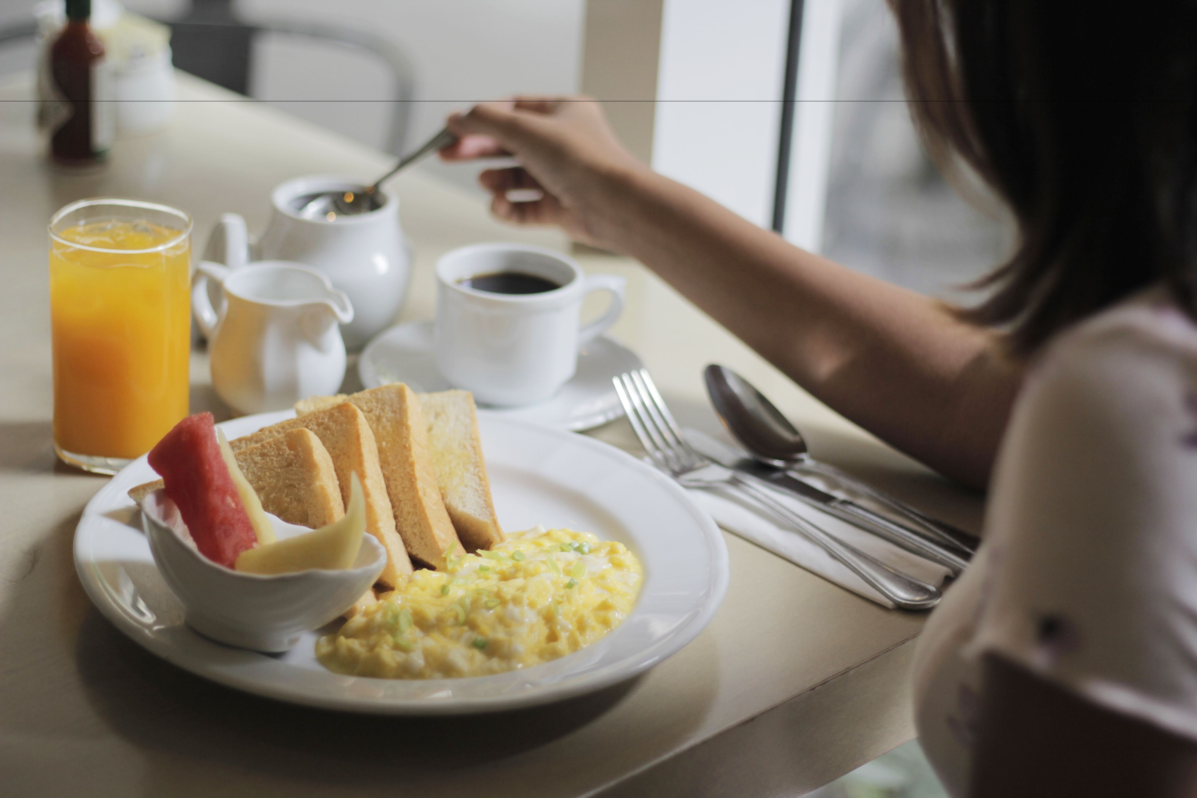 Uma mesa posta para o café da manhã, com torradas, ovos mexidos, uma tigela de frutas, um copo de suco de laranja e uma xícara de café.