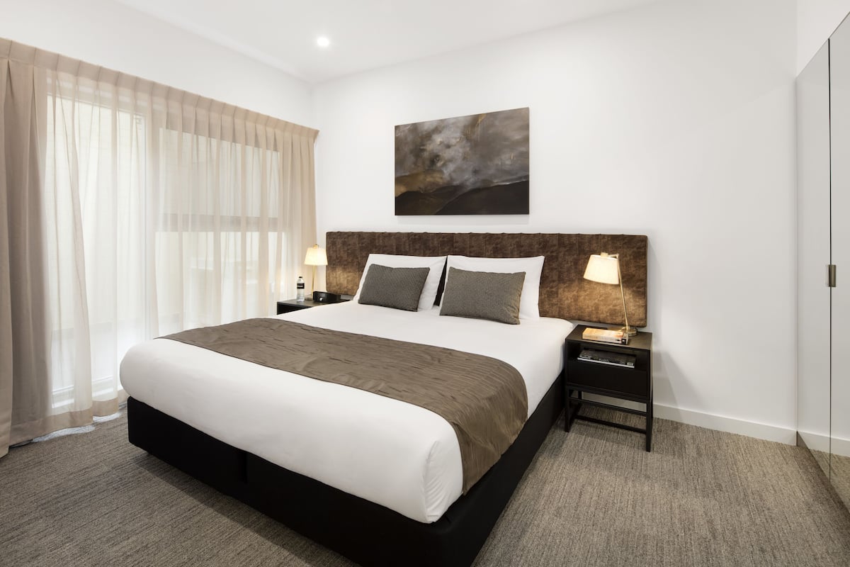 A modern hotel room with a large bed, a headboard with a painting, a bedside table with lamps, and sheer curtains.