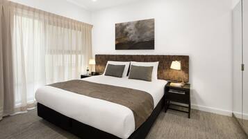 A modern hotel room with a large bed, a headboard with a painting, a bedside table with lamps, and sheer curtains.
