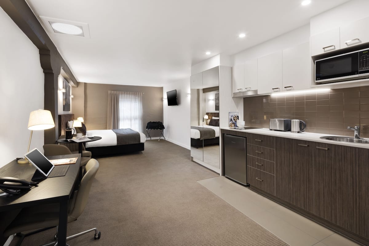 A modern hotel room with a kitchenette, a bed, a desk with a laptop, and a TV.