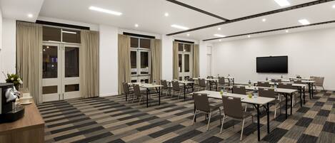 A conference room with multiple tables and chairs, a flat-screen TV on the wall, and a carpeted floor.