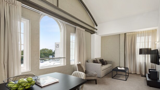 A modern hotel room with a large window, a sofa, a dining table with a bowl of apples, and a TV. - Quest Newcastle West (Newcastle West)