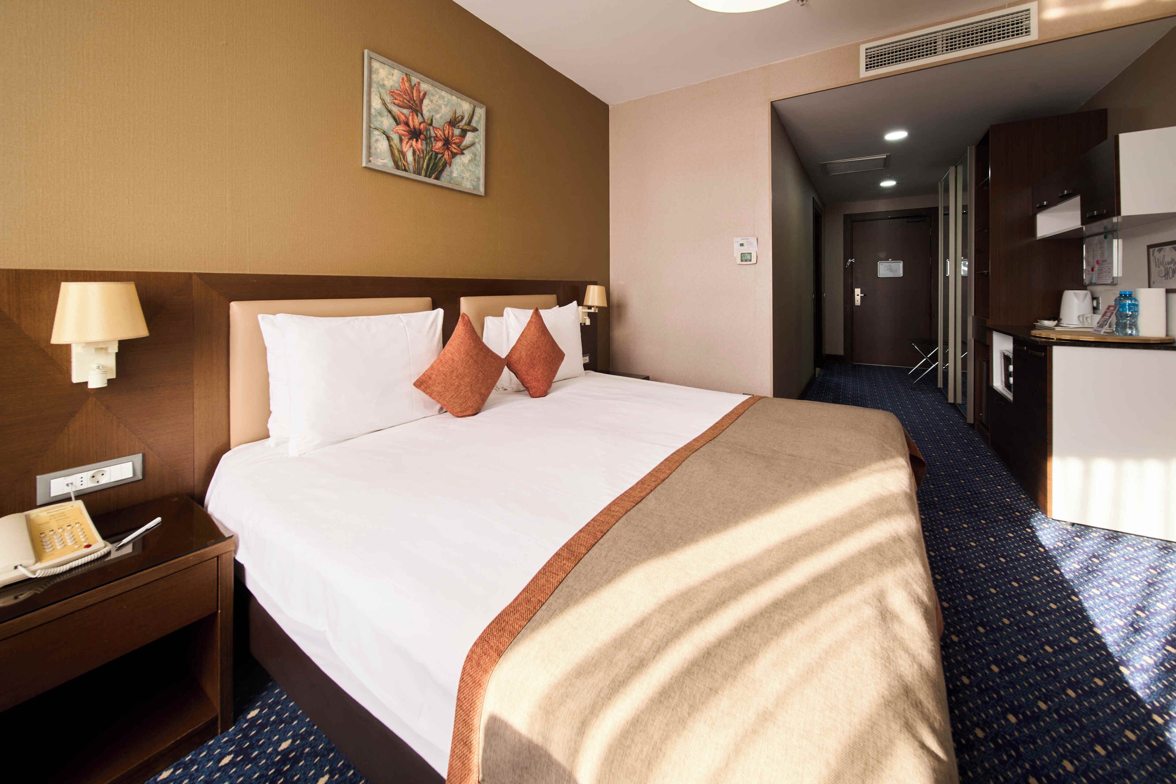 A hotel room with a bed, bedside tables, a painting, and a kitchenette area.