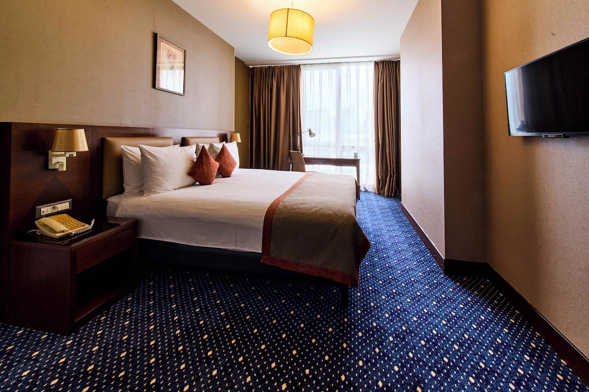 A hotel room with a bed, a desk with a lamp, a telephone, a TV, and a window with curtains.