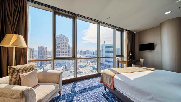 A hotel room with a large window, a bed, a sofa, a desk, and a TV. - Parkside Hotel & Apartments (Baku)