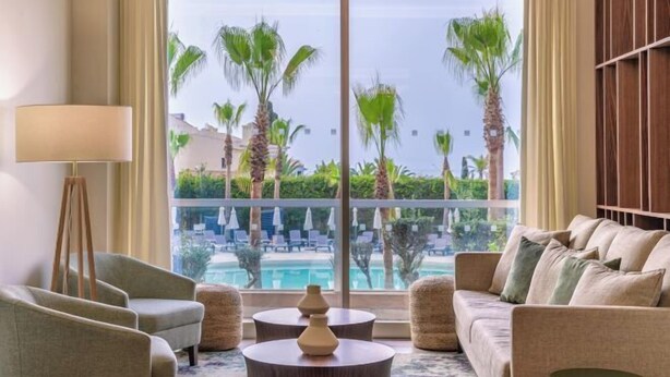 A hotel room with a large window overlooking a pool and palm trees. There is a sofa, a floor lamp, and a coffee table. - Sao Rafael Suites (Albufeira)