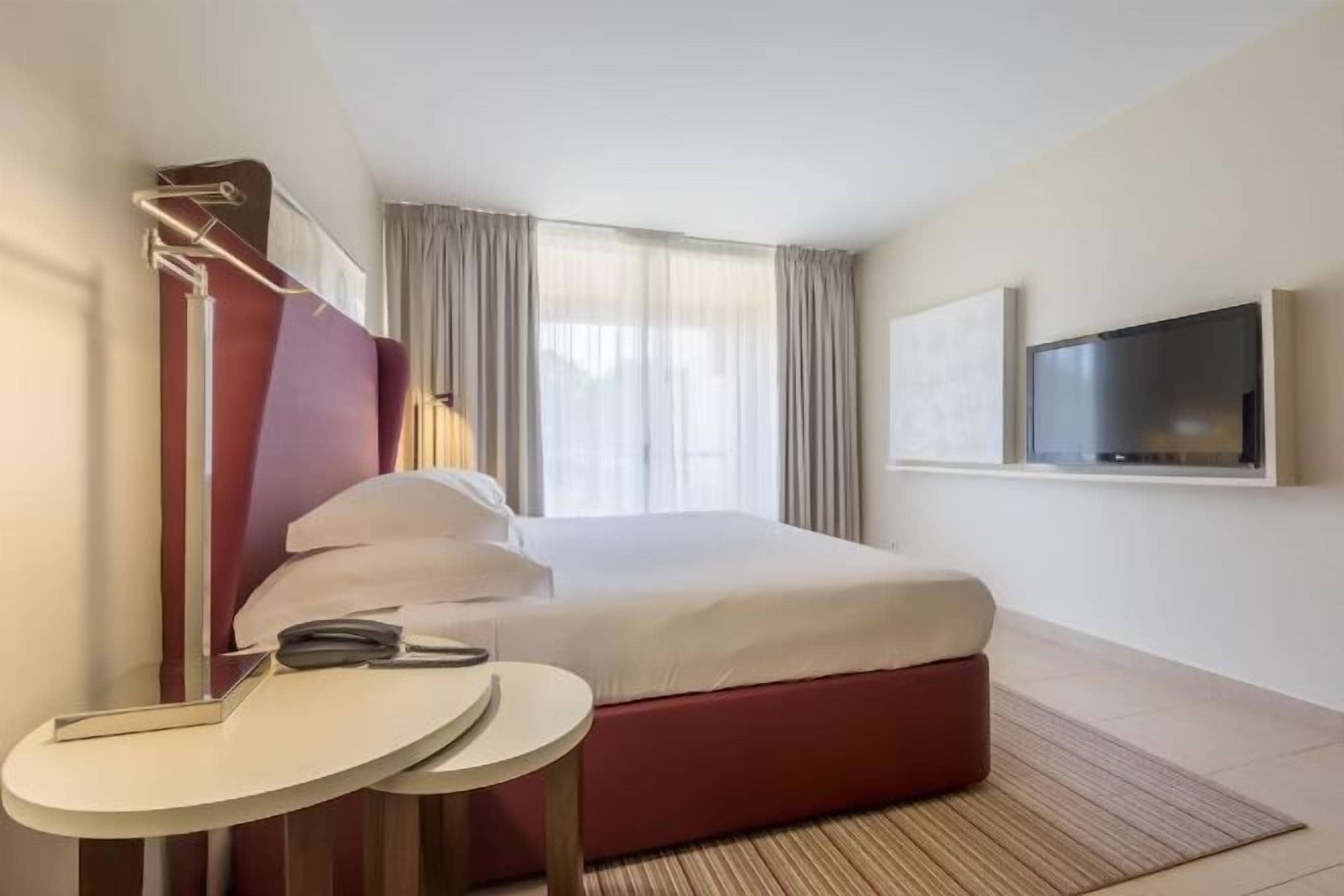 A hotel room with a bed, a TV mounted on the wall, a small round table, and a window with curtains.